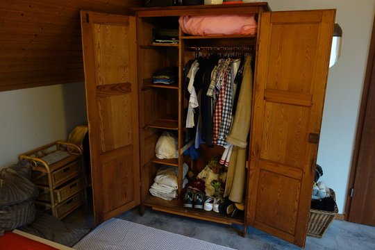 Unordentlicher alter h&ouml;lzerner Kleiderschrank mit Kleidung und Kinderpuppe. Ausmisten nach Kondo: Does it spark joy? Chaotischer Schrank aus Holz mit Klamotten und Gelumpe Chaotic Wardrobe Marie Kondo