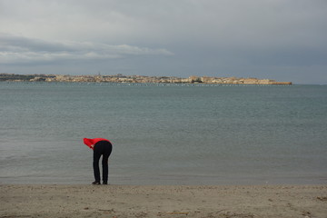 Popo am Strand mit Meer. Ass at the beach. Eine Person bückt sich nach etwas am Strand. Person an der Küste hebt einen Stein auf, um ihn ins Wasser zu schmeißen. Unvorteilhaftes Bücken. Knackarsch.  - obrazy, fototapety, plakaty