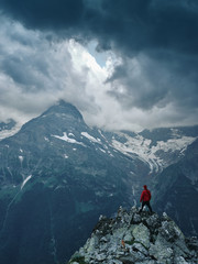 Alone hiker man in red jacket against the gloomy mountain top landscape with thunder cloudy sky,...