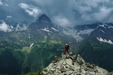 Obraz premium Alone hiker man with backpack and helmet against the gloomy mountain peak landscape with thunder cloudy sky, rocky ranges and peaks with glaciers. Extreme domestic travel and trekking. Local tourism.