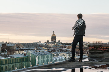 Fototapeta na wymiar A bearded guy in a leather vest and jeans stands on the edge of the roof with a view of St. Petersburg at sunset