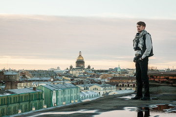 A bearded guy in a leather vest and jeans stands on the edge of the roof with a view of St. Petersburg at sunset
