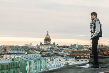 A bearded guy in a leather vest and jeans stands on the edge of the roof with a view of St. Petersburg at sunset