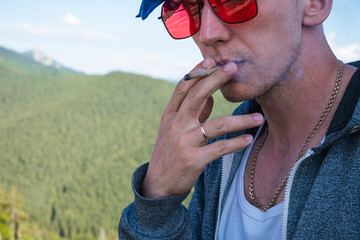 Close-up of young man smoking marijuana joint outdoors. Hemp blunt in the male hand. Cannabis is a...