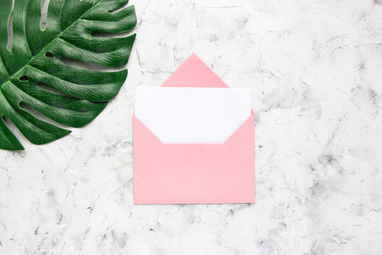 Pink envelope with white blank card and green monstera leaf on white concrete background. Flat lay, top view.
