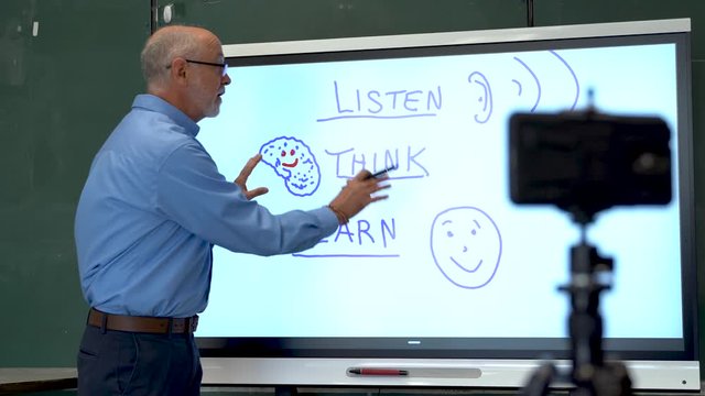 Portrait of very animated man using an interactive whiteboard in a classroom teaching to phone camera.