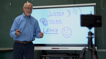 Portrait of very animated man using an interactive whiteboard in a classroom teaching to phone camera.