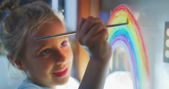 Authentic shot of a cute little creative happy smiling preschooler girl painter is drawing a rainbow on a glass of a window in a kitchen at home. Concept of happiness, arts, childhood, covid free