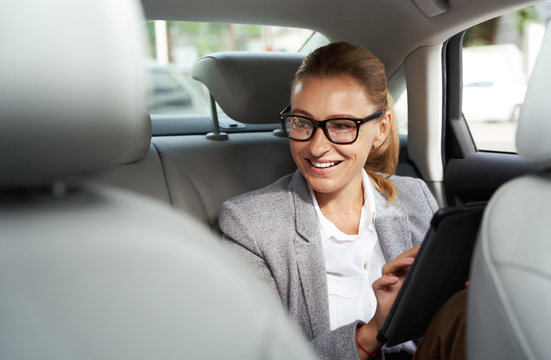 Working on the way to the office. Successful business woman wearing eyeglasses using digital tablet and smiling while sitting on back seat in the car