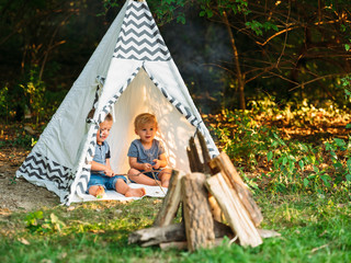 Two young boys playing in a backyard teepee campsite under the summer sunset