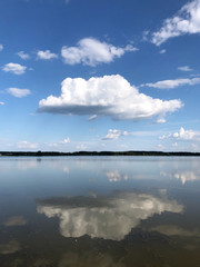 Naklejka na ściany i meble White big fluffy cloud on a blue sky reflected in a clear lake water with a forest horizon line. Summertime sunny day peaceful relaxing concept