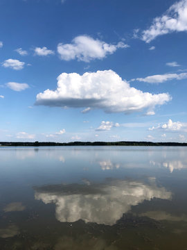 White big fluffy cloud on a blue sky reflected in a clear lake water with a forest horizon line. Summertime sunny day peaceful relaxing concept © Nastassia Kudzina