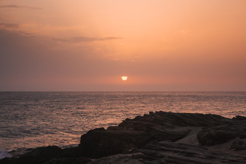 Beautiful sunset on the sea with the sun going down on the horizon and rocks in the foreground.