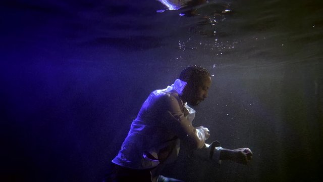 A close-up of a muscular African-American man is under water at a depth of, around him, darkness and bubbles, he is in an unbuttoned white shirt with a bare chest, making movements with his hands.