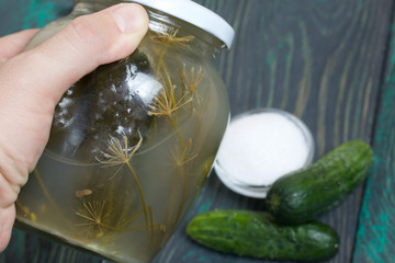 A jar of pickled cucumbers in a man's hand. Nearby are fresh cucumbers and a container of salt.