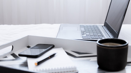 Montando la  oficina desde la comodidad del hogar. Laptop junto a una bandeja con una taza de café un teléfono móvil y un anotador sobre una cama de sábanas blancas en una luminosa habitación.