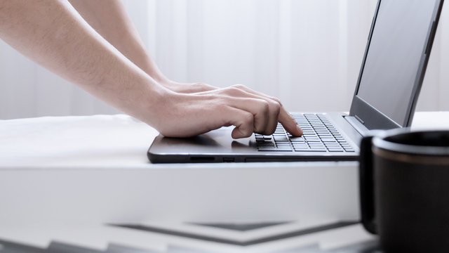 Trabajando en el ordenador port&aacute;til desde la comodidad del hogar. Primer plano de manos cauc&aacute;sicas de hombre escribiendo sobr eun teclado negro de una laptop en una habitaci&oacute;n blanca e iluminada