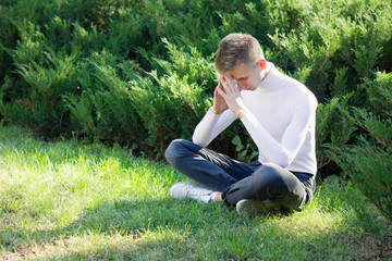 Naklejka na ściany i meble A young blond guy, dressed in a white jacket and black jeans, sits on the ground, clasping his legs and hands together, against the background of nature.
