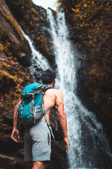 Obraz premium Travel and freedom. Young shirtless boy in shorts with rucksack enjoying tropical waterfall view.