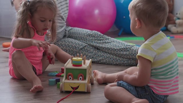 Childhood, games, upbringing and development concept - general plan of small children boy and girl of same age with mom are playing with children's wooden car constructor and puzzles in kid playroom