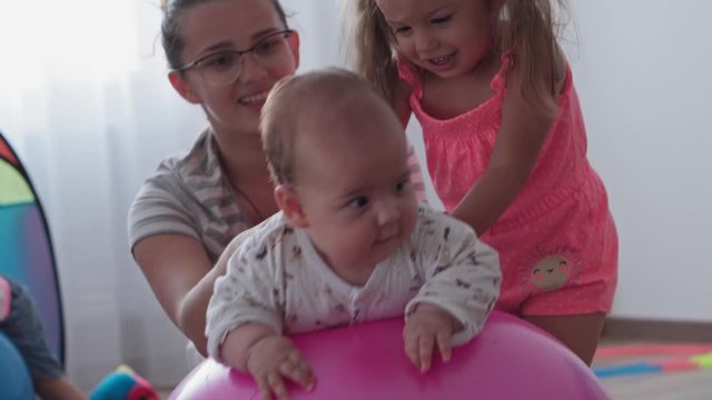 Childhood, Motherhood, games, upbringing and development concept - close-up young mom are joyfully doing exercise and masah with two newborn and little daughters on fitball and smiling in kid playroom
