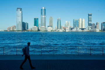 Fototapeta na wymiar New York, NY / USA - 8/20/20: an image of a commuter walking on the Battery Park City Esplanade with the Hudson River and the Jersey City skyline behind him