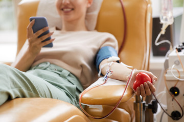 Close-up of young woman lying on the couch with mobile phone while donating blood at the laboratory - obrazy, fototapety, plakaty