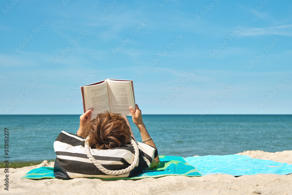 Wall mural Caucasian woman lying on the beach reading a book on bright summer day