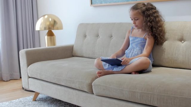 Cute little girl preschool child using a digital tablet technology device looks telephone.Plays in the phone sitting on the sofa alone.Children tech addiction concept play game at home.