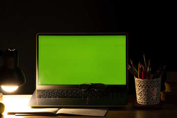laptop with green screen on the table with books near the included bright lamp in the dark bedroom