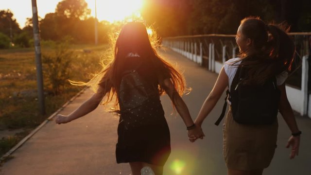 Two friends schoolgirls teenagers with backpacks run down the street returning from school at sunset