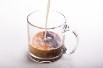 Cream poured in Black Coffee into glass mug on white surface
