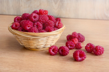Small wicker basket full of ripe raspberries stands on wooden table. Several berries lies on the table next to the basket. Selective focus. Delicious organic food theme.