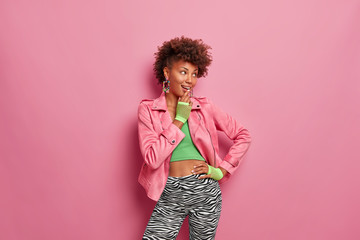 Studio shot of fashionable dark skinned girlfriend looks aside with cheerful pensive look, keeps hand on hip, dressed in stylish sport clothes, isolated over pink background. Fashion concept - obrazy, fototapety, plakaty