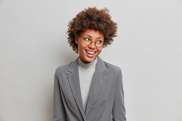 Cheerful prosperous businesswoman in elegant grey formal clothes, gazes happily aside, wears transparent glasses, feels successful to lead new project, accomplished business affairs, owns company