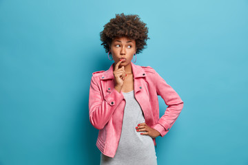 Half length shot of pensive young female model with curly hair, keeps index finger on lips, looks thoughtfully aside, ponders over right decision, wears grey dress and pink jacket, recalls something