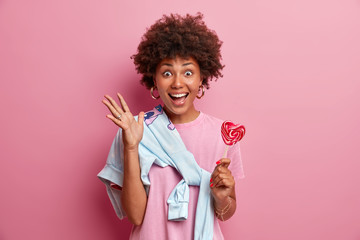 Overjoyed Afro American teenage girl glad to be in merry company, raises hand, holds sweet candy, wears sweater tied over shoulder, smiles broadly, isolated on pink background. Youth, leisure concept