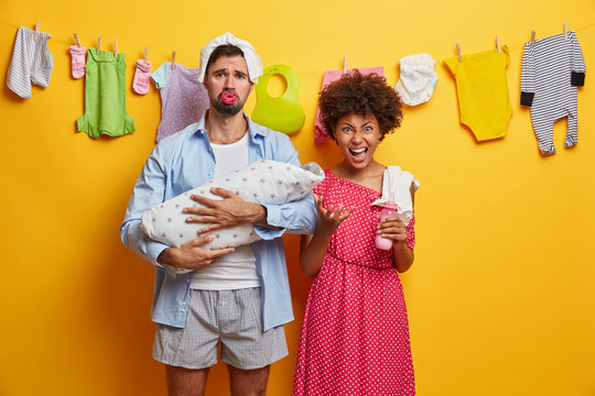 Small Newborn Daughter Sleeps Comfortably In Fathers Embrace. Tired Couple Care About Infant. Upset Dad With Diaper On Head, Holds Baby Wrapped With Blanket. Irritated Mother Going To Feed Kid