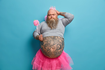 Fathers day holiday. Serious funny dad with big belly, wears princess outfit, holds magic wand, receieves congratulations from children, isolated over blue background. Playful male fairy indoor