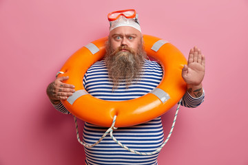 Serious stout man has thick beard makes no gesture, has safety beach holiday, poses with inflated lifering, wears striped sailor shirt, isolated on pink background. Swimming time, summer concept