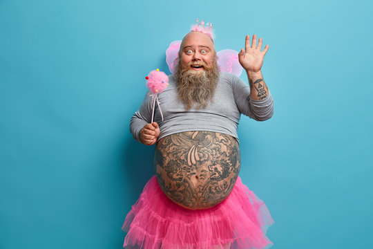 Horizontal positive man animator on childrens party, dressed in fairy costume, waves palm in hello gesture, has good mood during holiday, being kind magician, has big tattooed belly, blue background