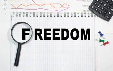 freedom in a magnifying glass on a notepad against the background of graphs, calculator and paper clips.