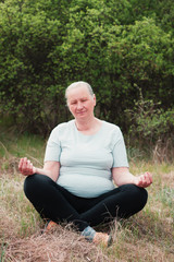 Naklejka na ściany i meble Portrait of a senior woman, wearing a white T-shirt and black pants, meditates in nature. Meditation, yoga and relaxation concept.