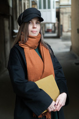 Long-haired girl student in a black coat with a vintage cap and a red scarf, posing with a book in a shabby quiet courtyard