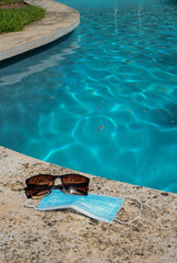 Blue face mask for protection against the Corona Virus (COVID-19), sunglasses laying beside a swimming pool during coronavirus worldwide pandemic holidays