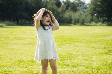 Obraz premium Little Hispanic girl smiling and playing in a natural park - little girl in a white dress and hat in the middle of a green field - cute little girl playing with her hat.