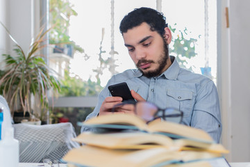 student searches for content on her mobile, behind a stack of open textbooks on her desk, studying...