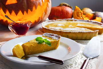 Piece of homemade halloween pumpkin cake with leaf of mint on white plate. Jack-o-lantern, fork and cowberry jam around