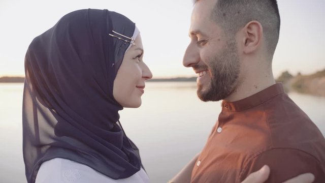 Muslim love story close-up video in 4k. Mixed couple embraces, caress and looks at sunset on the lake . Woman weared in hijab looks to her man. Advert for on-line dating agency