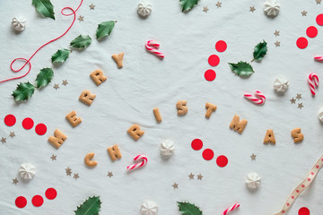 Eco-friendly Xmas decor with natural holly leaves. Flat lay on white textile. Merry Christmas text made of cookies. Xmas decor silhouettes cut out from brown and red paper and sweet marshmallows.